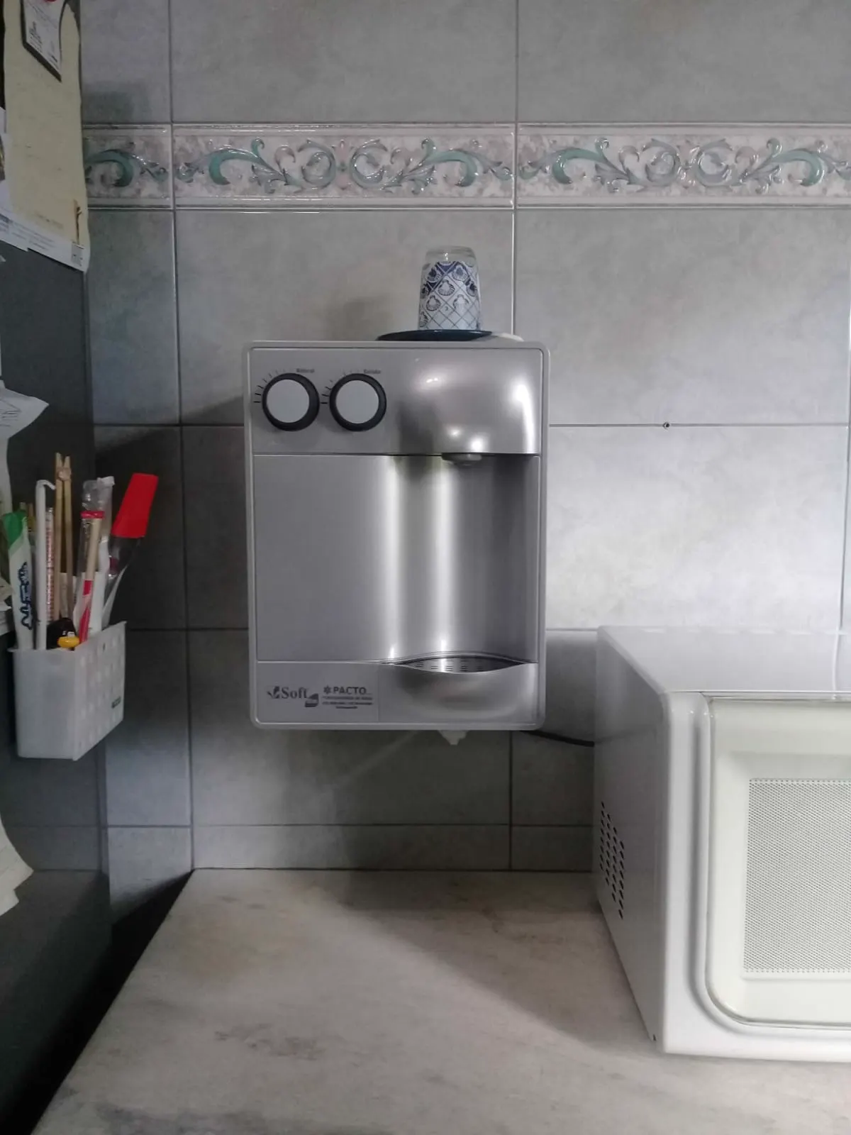 Purificador de água prata instalado em parede de azulejos ao lado de um micro-ondas